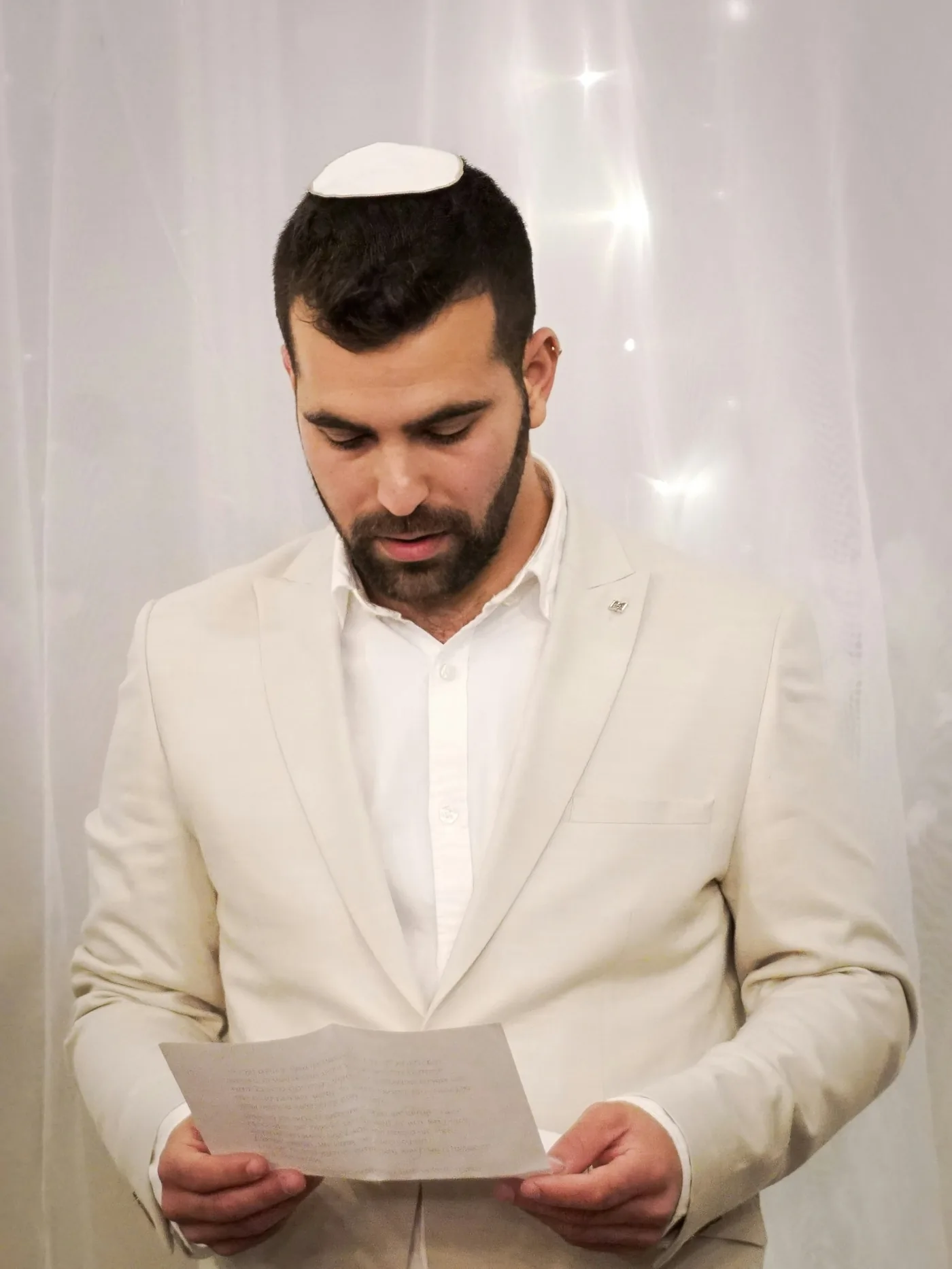 A groom in a white suit and kippah reading from a page before his wedding.